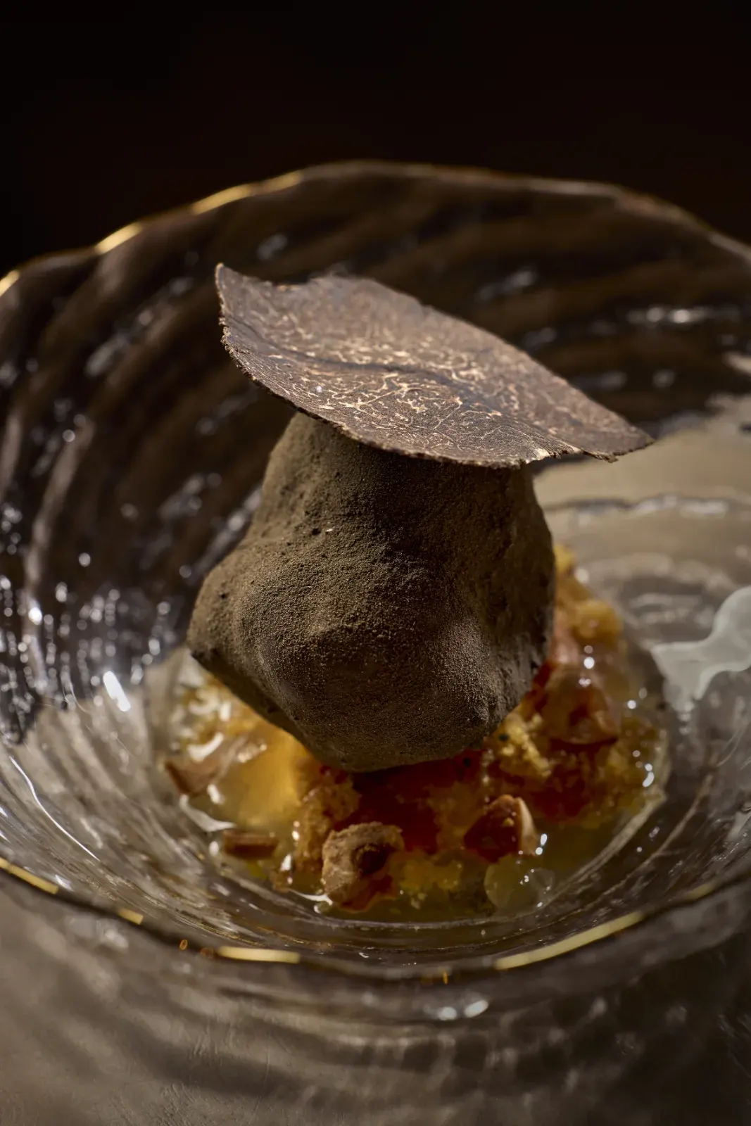 Bocado coronado con una lámina de trufa, sobre un crumble dorado y su jugo, en cuenco de cristal